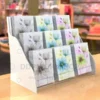 Three-tier white Perspex greeting card display stand holding A5 floral cards, placed on a retail countertop.