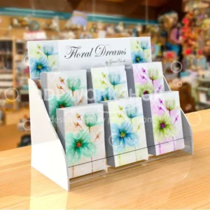 White acrylic two-tier card display rack with clear acrylic shelves showcasing floral greeting cards, set on a wooden countertop in a retail shop.
