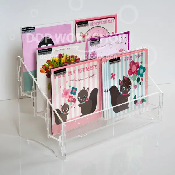 Clear acrylic three-tier card display stand filled with colourful square greeting cards, shown on a white surface.