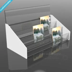 A tiered acrylic coaster display stand with clear stepped shelves and white side panels, showcasing square coasters with scenic artwork.