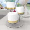 Three-tier clear acrylic cake stand displaying elegant white cakes with gold ribbon, arranged in a cascading waterfall effect.