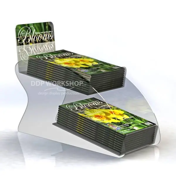 Clear acrylic two-tier counter display stand holding stacks of "Blooms" magazines with curved side panels.