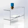 Clear acrylic display table with integrated shelf, shown with an open book and decorative vase on a glossy white background.