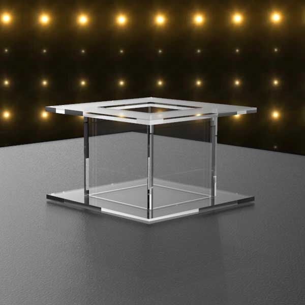 Clear acrylic square cake separator with open fillable center, polished edges, and tiered design, displayed against a black backdrop with golden lights.