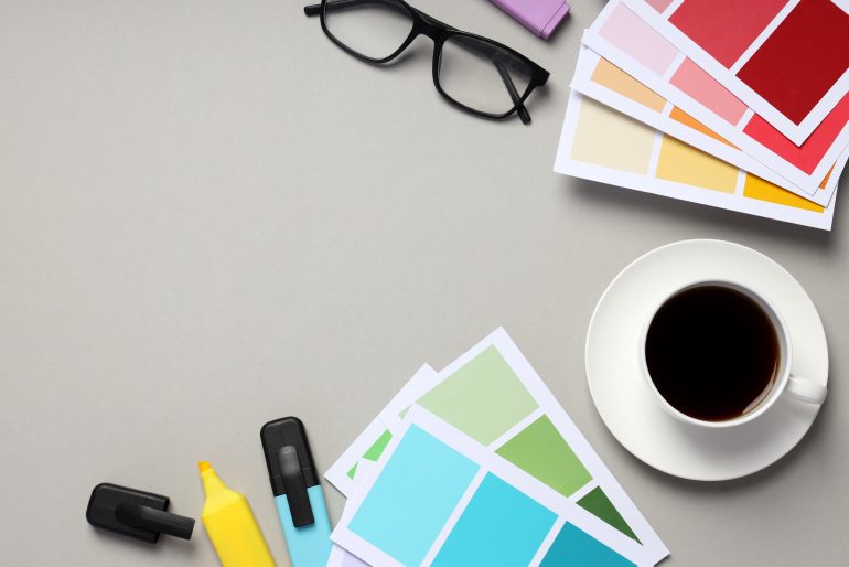 A workspace with colour swatches, highlighters, eyeglasses, and a cup of coffee, representing the design and artwork process.